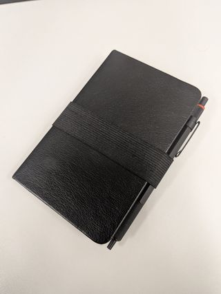 A photo of my notebook. A simple, black hardcover book. A black elastic band holds a black pen to the side. The pen is a Rotring 600