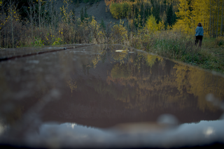 A photograph taken just before the sun made it over the mountains. In the foreground, a picnic table is covered in a thin film of water, reflecting the background. The trees of Guanella Pass are visible both in the reflection and above the table in the frame. Aspen and pine trees in contrasting shades of green and gold litter the mountainside. In the far background, a woman stands off to the side facing away from the camera. She's wearing a denim jacket and black leggings.