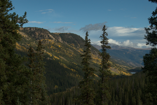 A photo from further up Guanella pass, looking down into the valley. Otter Peak is on the left side of the frame, with groves of aspens interspersed among the pines, their bright yellow leaves shining brightly in the morning sun. Two skinny pine trees highlight the center of the frame, much closer to the camera than the mountain in the background. Two more pines line the left and right edges of the frame, even closer to the camera. Off in the distance, in the right half of the frame, Clear Lake can be seen poking out between the mountain slopes.
